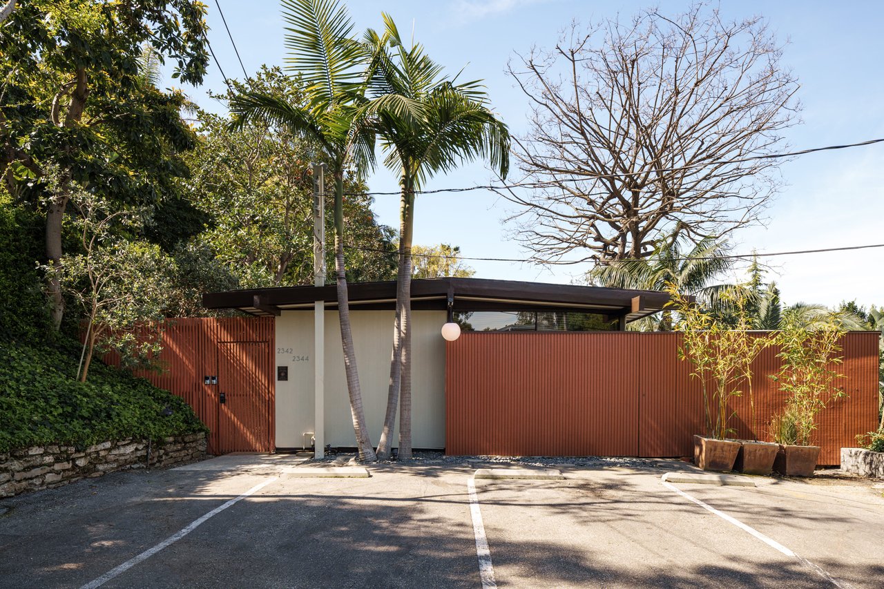 Modern building entrance behind a rust-red slatted fence, framed by palm trees.