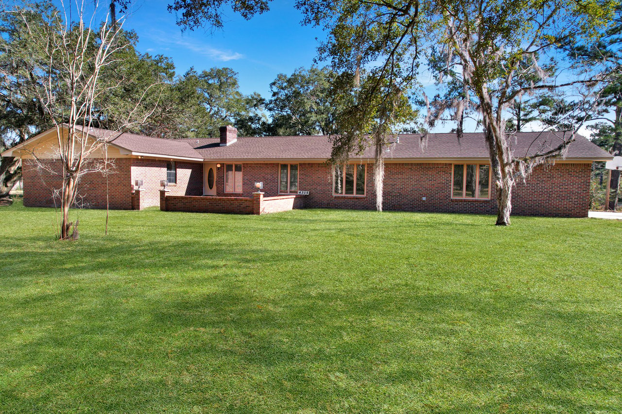 Single-story brick house with large front lawn, surrounded by trees under a clear blue sky. The scene conveys tranquility and suburban charm.