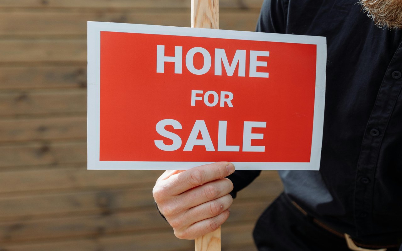 A person holds a red-and-white HOME FOR SALE sign.