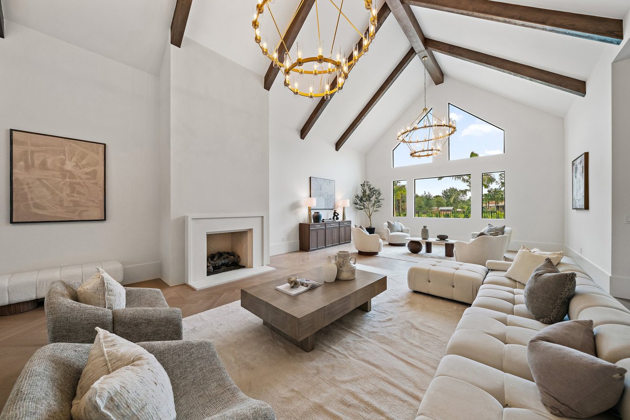 Vaulted living room with exposed beams, gold chandeliers, a fireplace, and windows overlooking greenery.