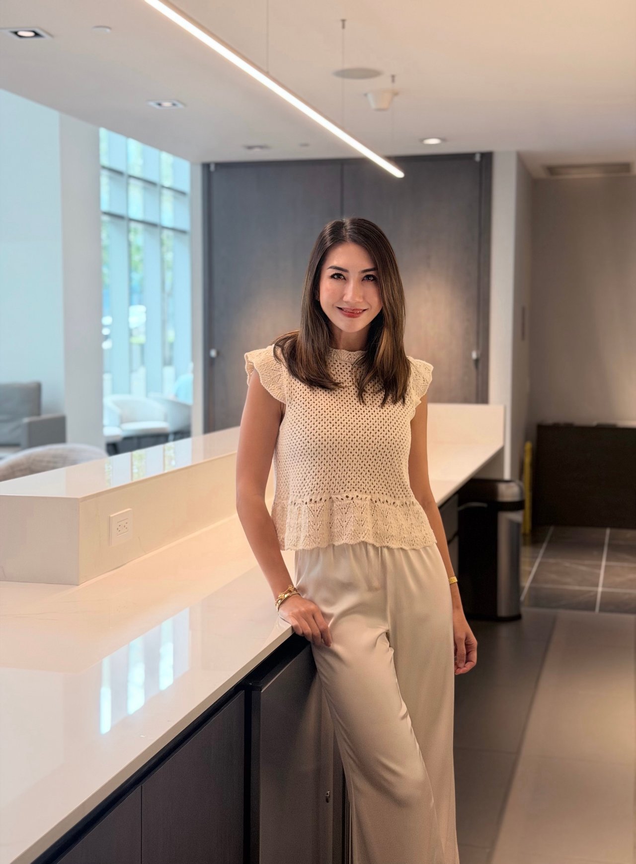 Leona Li stands beside a white counter in a modern office, wearing a cream knit top and light trousers.