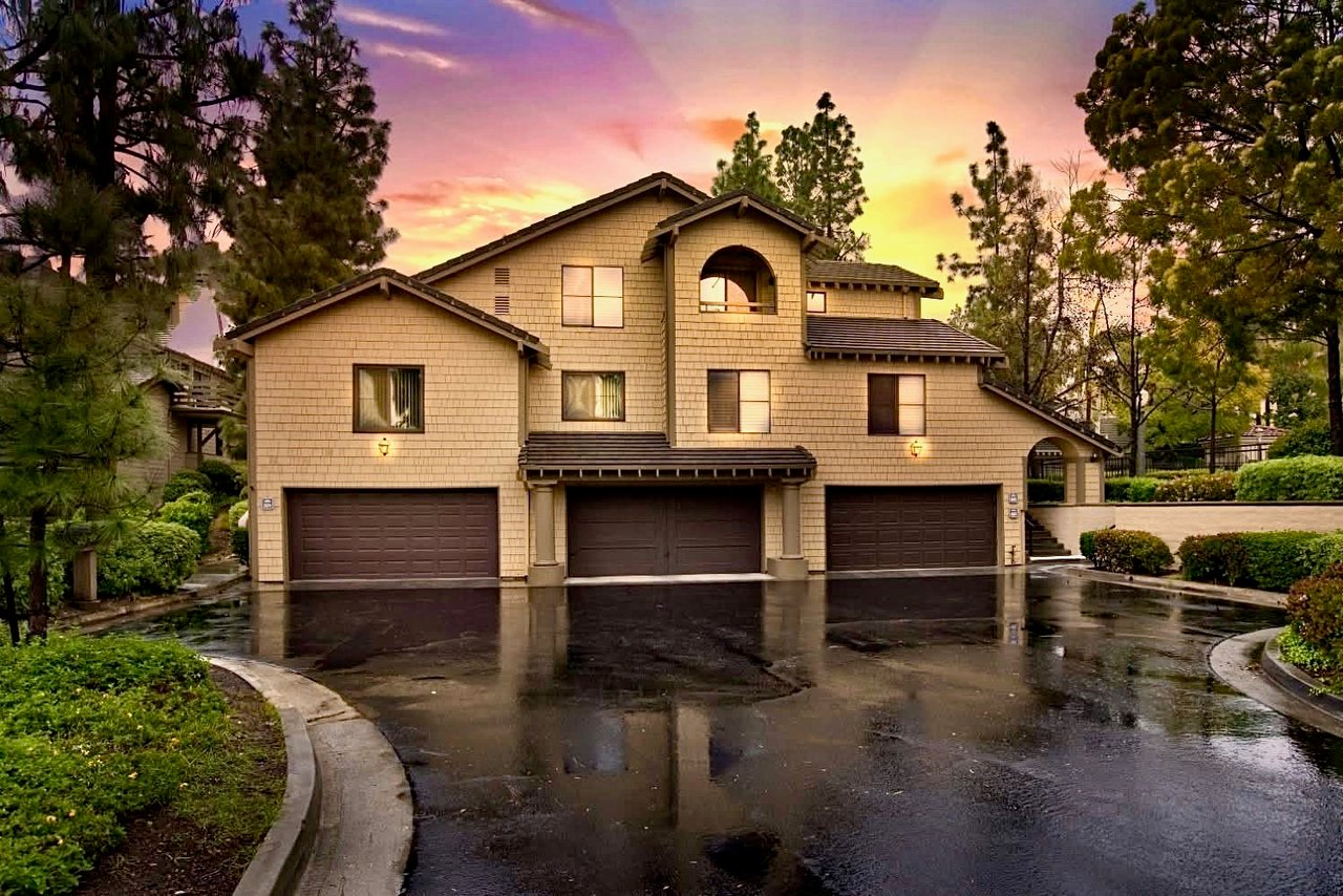 Two-story residential building with three garage doors and a wet reflective driveway at sunset.