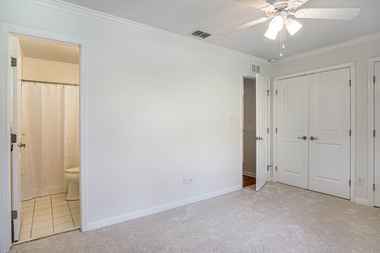 Empty room with beige carpet and white walls. A ceiling fan with lights hangs from the ceiling. Open door reveals adjacent room, another to a bathroom.
