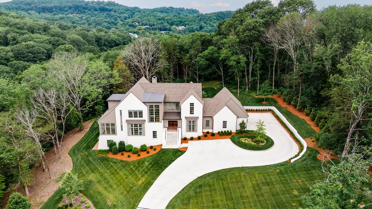 Aerial view of a white multi-gabled house with a curved driveway and wooded surroundings.