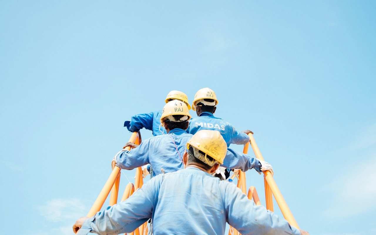 Four construction workers climb an orange ladder, seen from behind against a clear blue sky.
