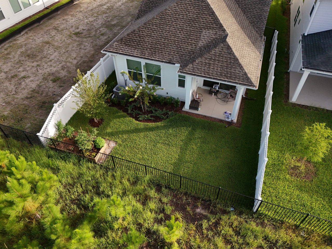 Aerial view of a home’s fenced backyard, landscaped garden beds, lawn, and covered patio.