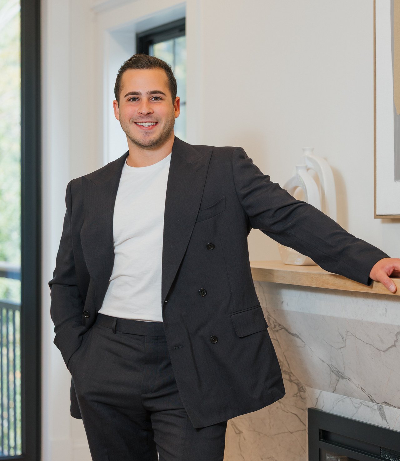 Alex MacDonald in a dark suit jacket and white shirt, leaning against a marble fireplace.