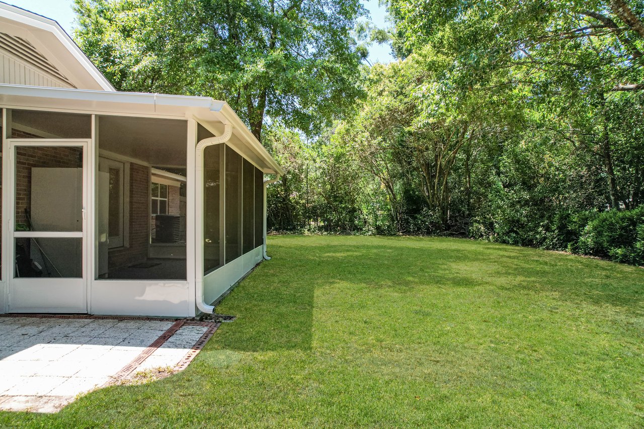 Screened patio attached to a house, surrounded by a grassy yard and bordered by lush green trees. Sunlight creates soft shadows, conveying a peaceful, suburban setting.
