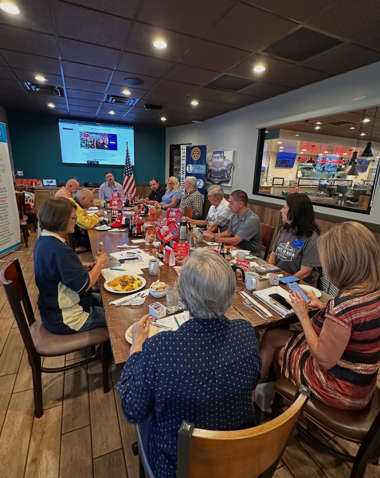 Adults share breakfast around a long table in a restaurant meeting room, with an American flag and Rotary banner.