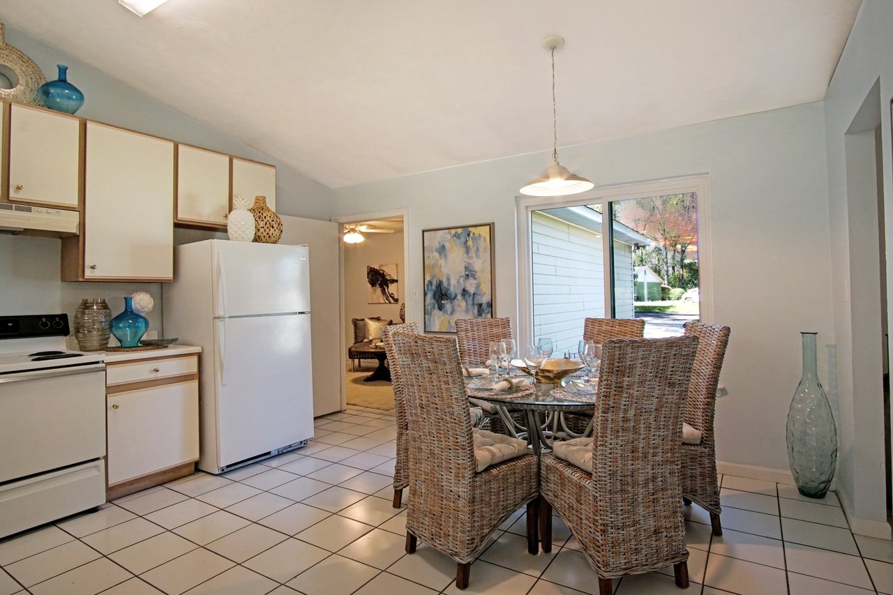Cozy kitchen with tiled floor, wicker chairs around a glass-top dining table, and soft lighting. Warm, inviting atmosphere with abstract wall art.