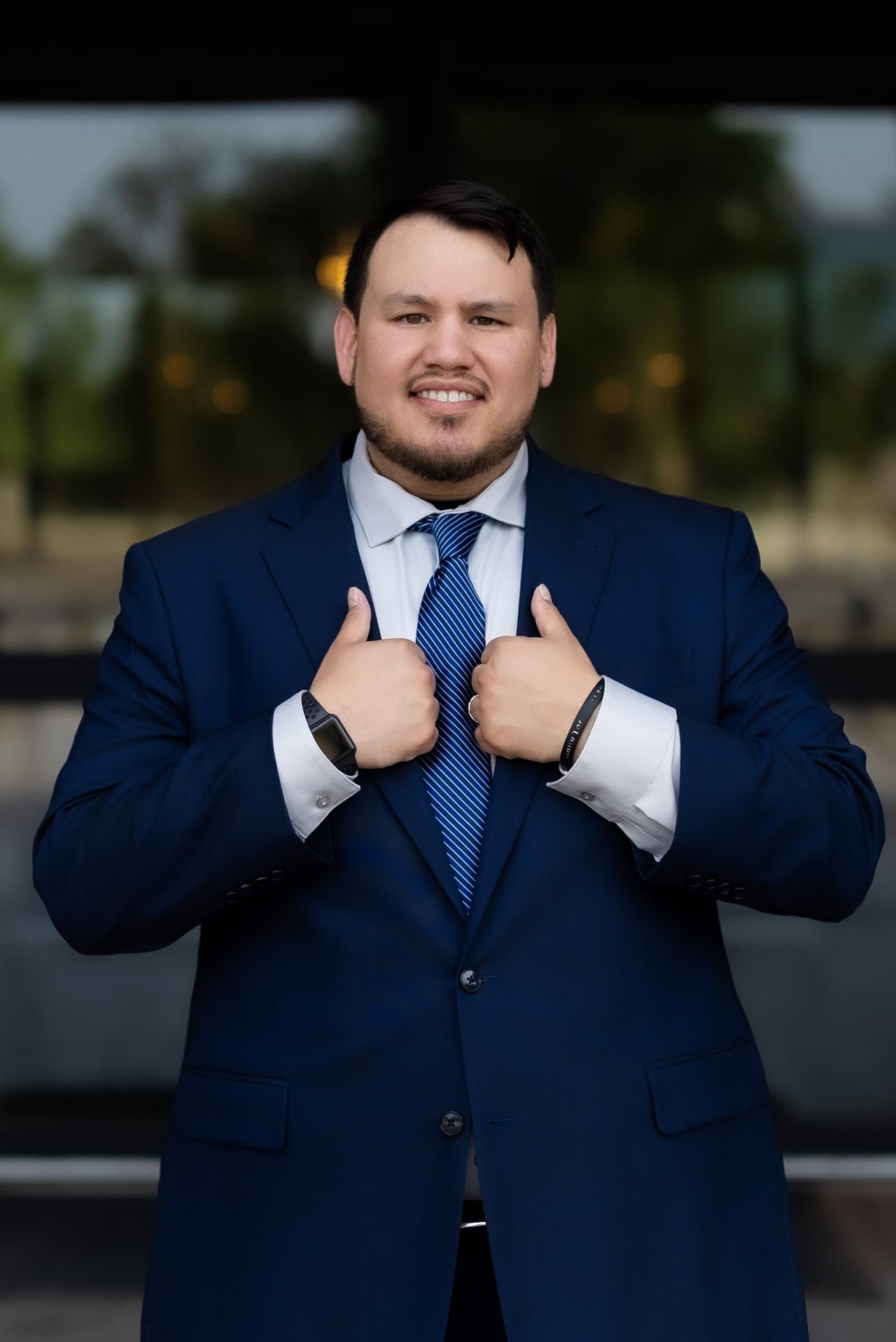 Bobby Nunez in a navy suit and striped tie, adjusting his lapels in front of a building entrance.