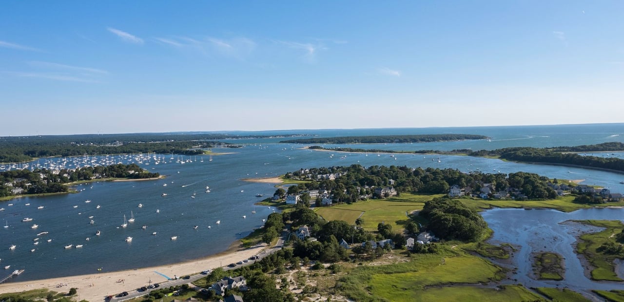Aerial view of a Cape Cod harbor with moored boats, shoreline homes, and tidal marsh.