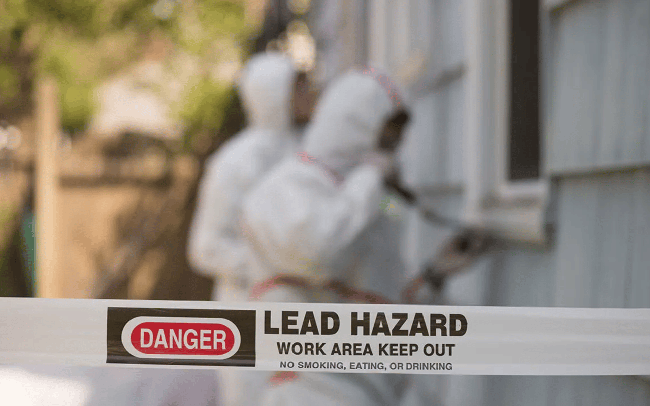 Lead hazard warning tape blocks an outdoor work area where a worker in protective coveralls is working.