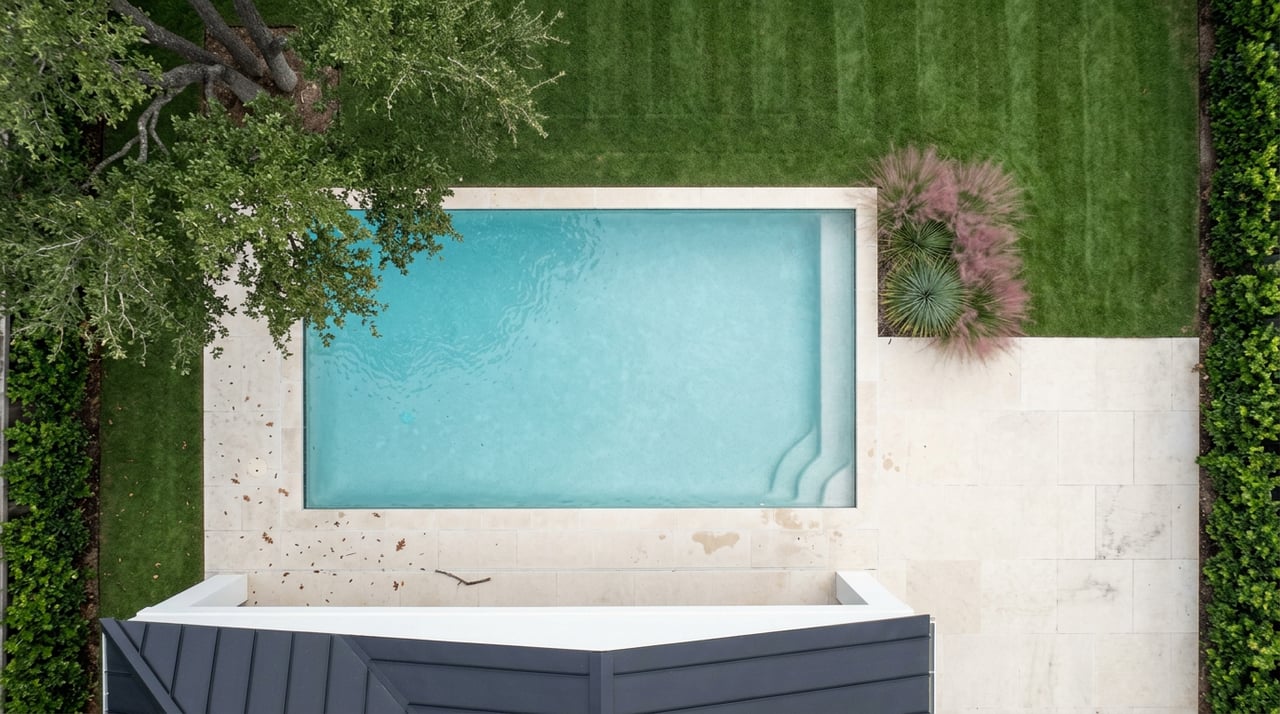 Overhead view of a rectangular pool bordered by pale limestone decking, striped lawn, and a live oak canopy.