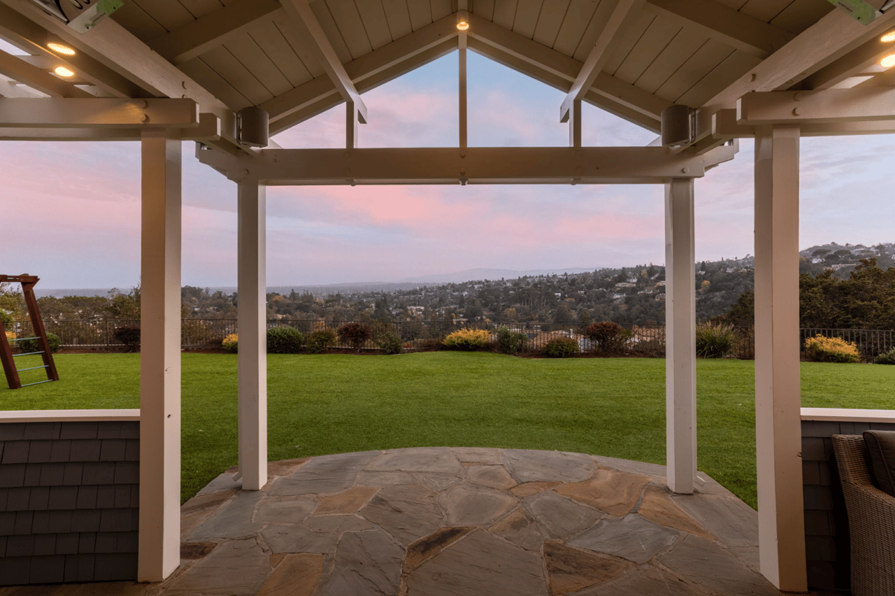 Covered stone patio and pergola overlooking a green lawn and residential hills at dusk.