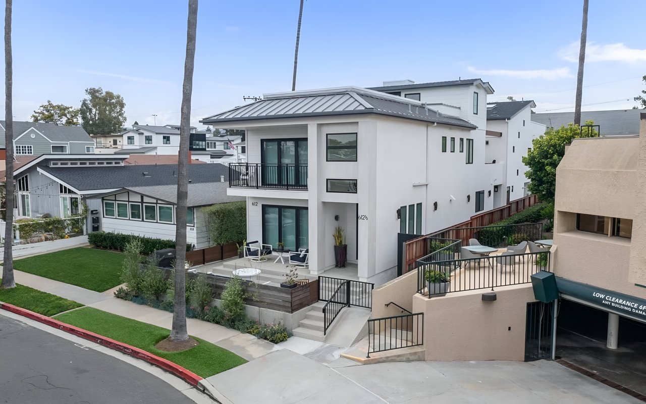  An aerial view of a modern, multi-story white house with a flat roof, balconies, and a patio, surrounded by palm trees.