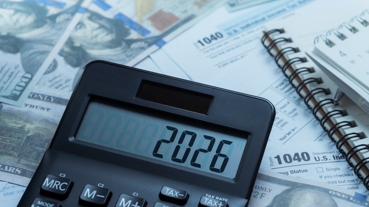 Calculator displaying 2026 on tax forms beside cash and a spiral notebook.
