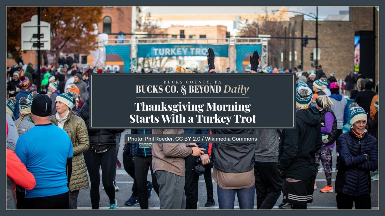 Runners in winter hats and jackets gather on a city street under a Turkey Trot banner at the 2021 Des Moines Turkey Trot.
