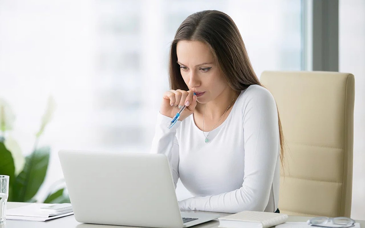Woman thoughtfully looking at a laptop, holding a pen near her lips.