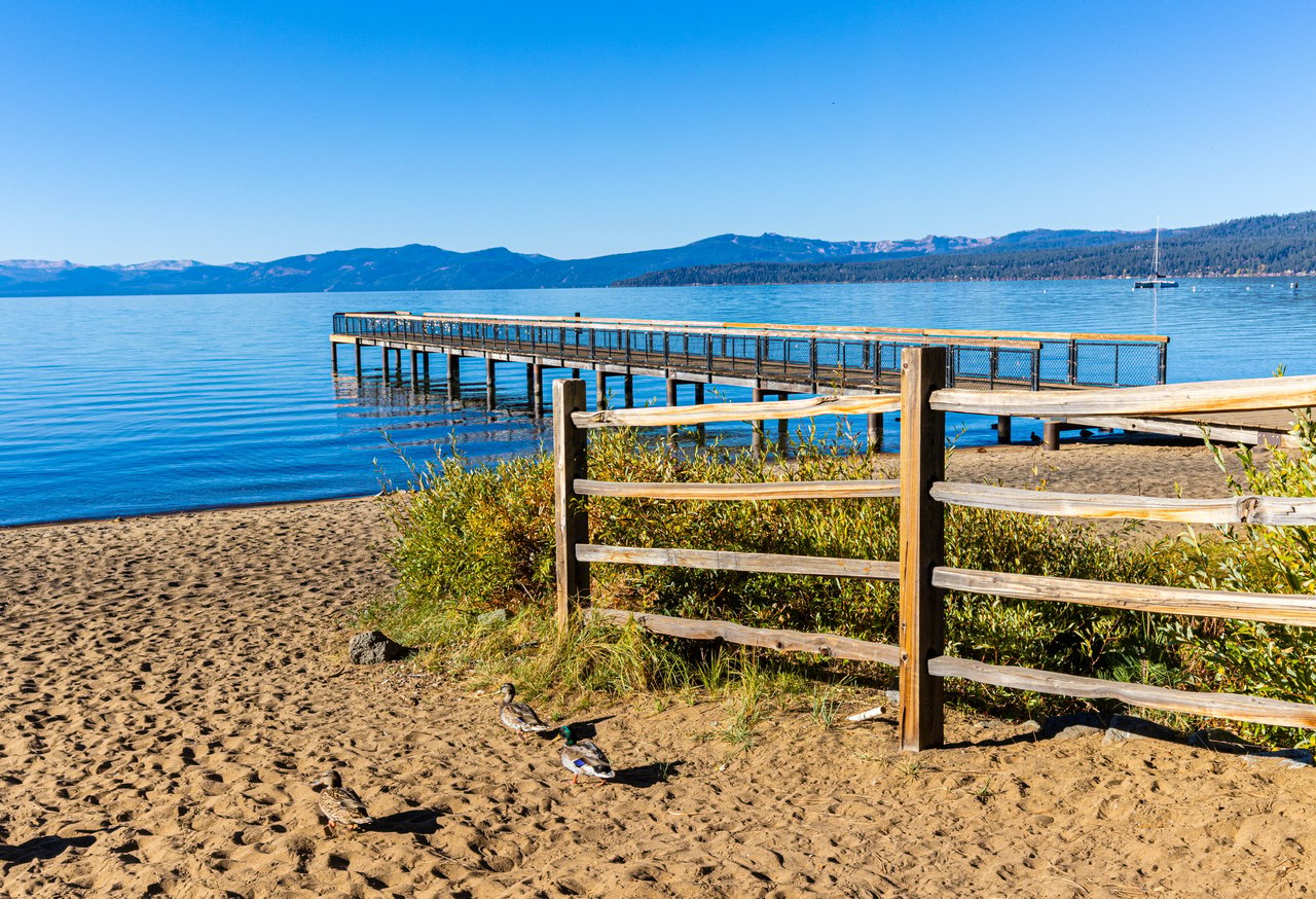 Luxury lakefront home on Lake Tahoe with a private pier extending into clear blue water, framed by pine trees and the Sierra Nevada mountains
