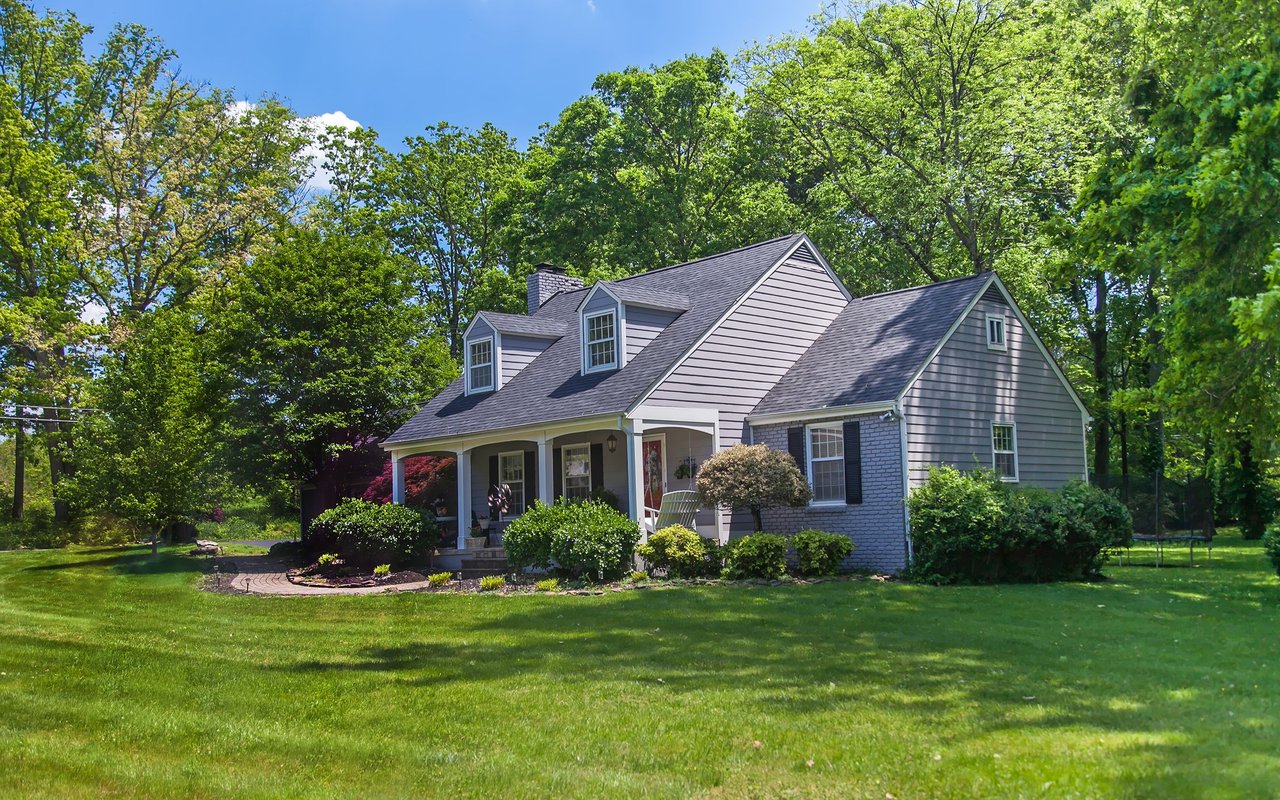 Gray two-story house with dormer windows and a covered porch, set among mature trees and a broad lawn.