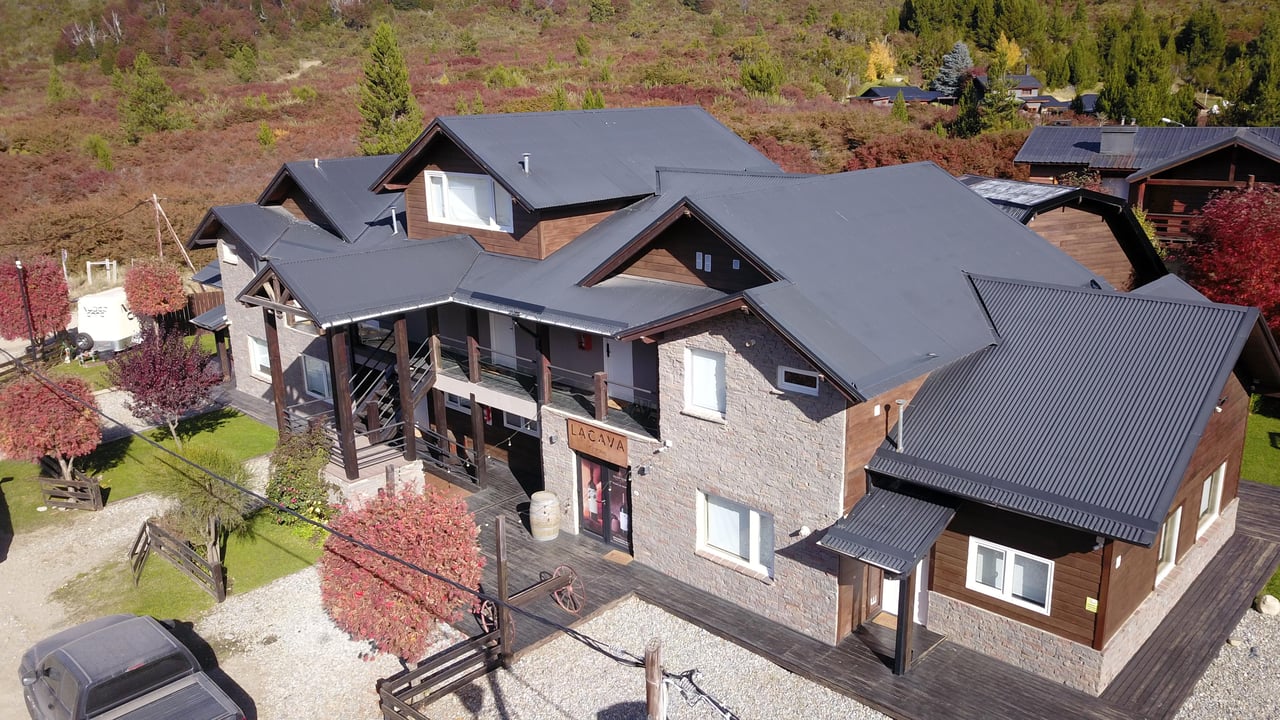 Stone-and-wood lodge with multiple dark roofs and balconies, viewed from above against an autumn hillside.