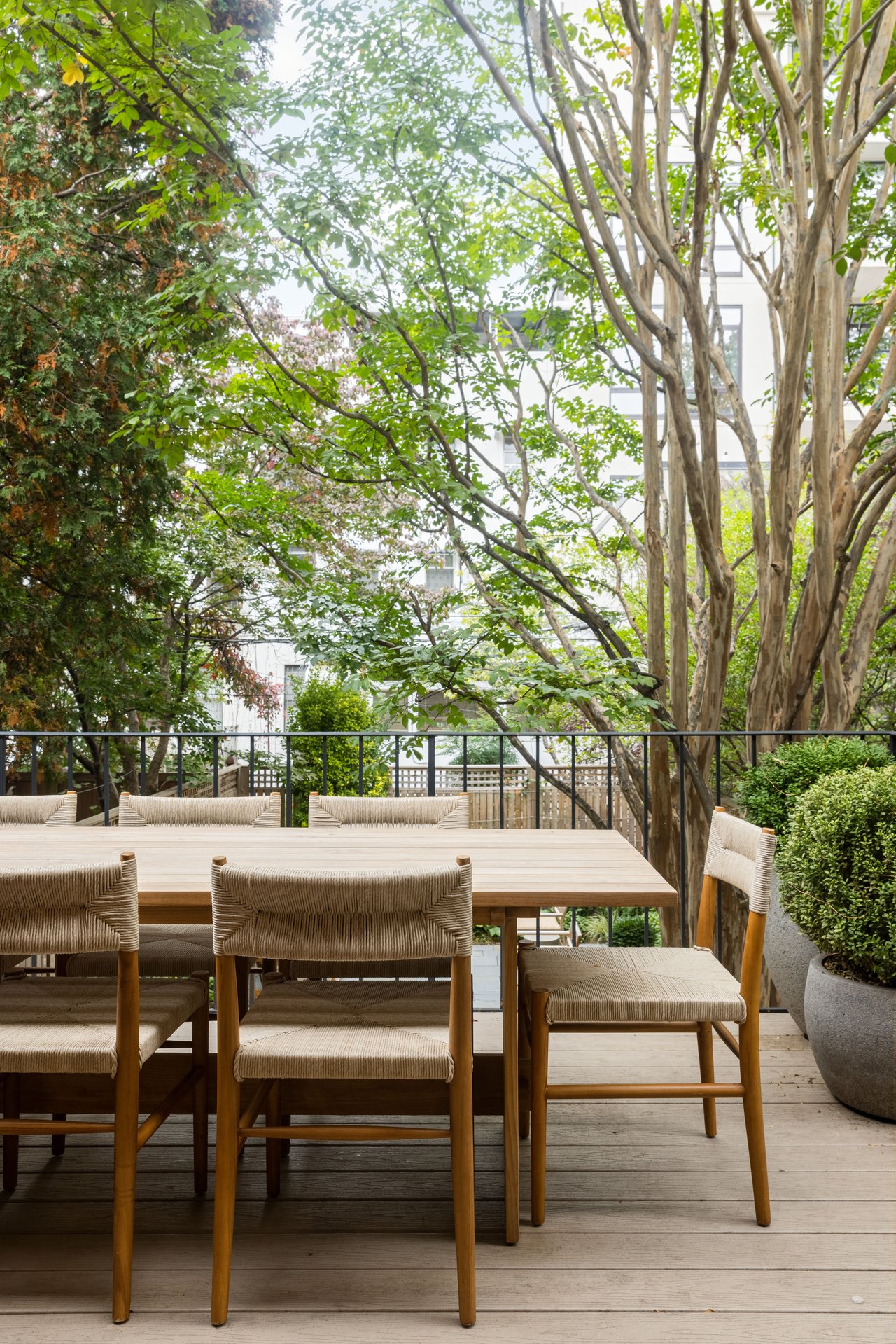 Close-up of an outdoor dining area featuring a light wood table and woven rope chairs, set on a deck surrounded by lush trees and potted greenery.