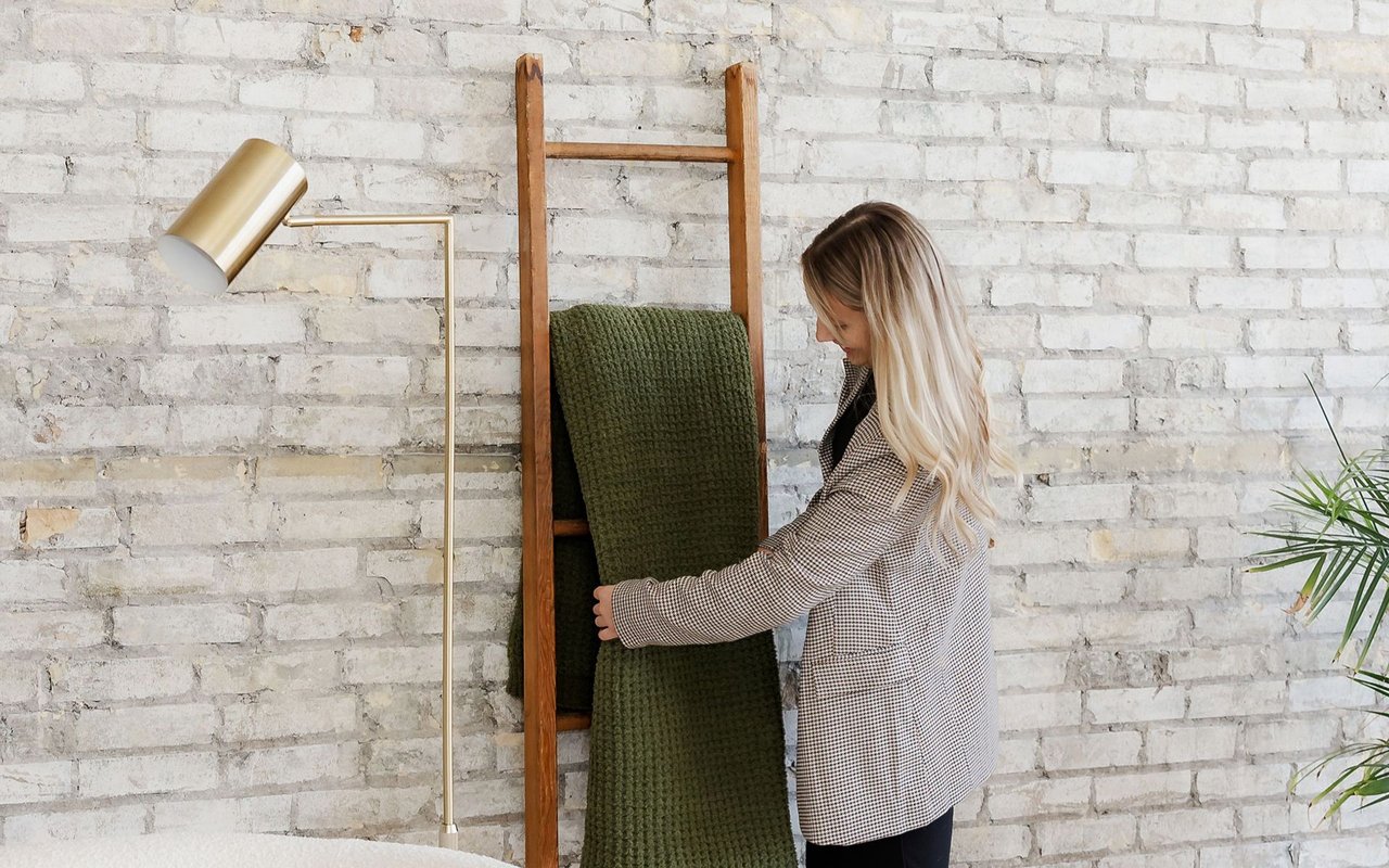 Woman arranging a dark green blanket on a wooden ladder beside a whitewashed brick wall.