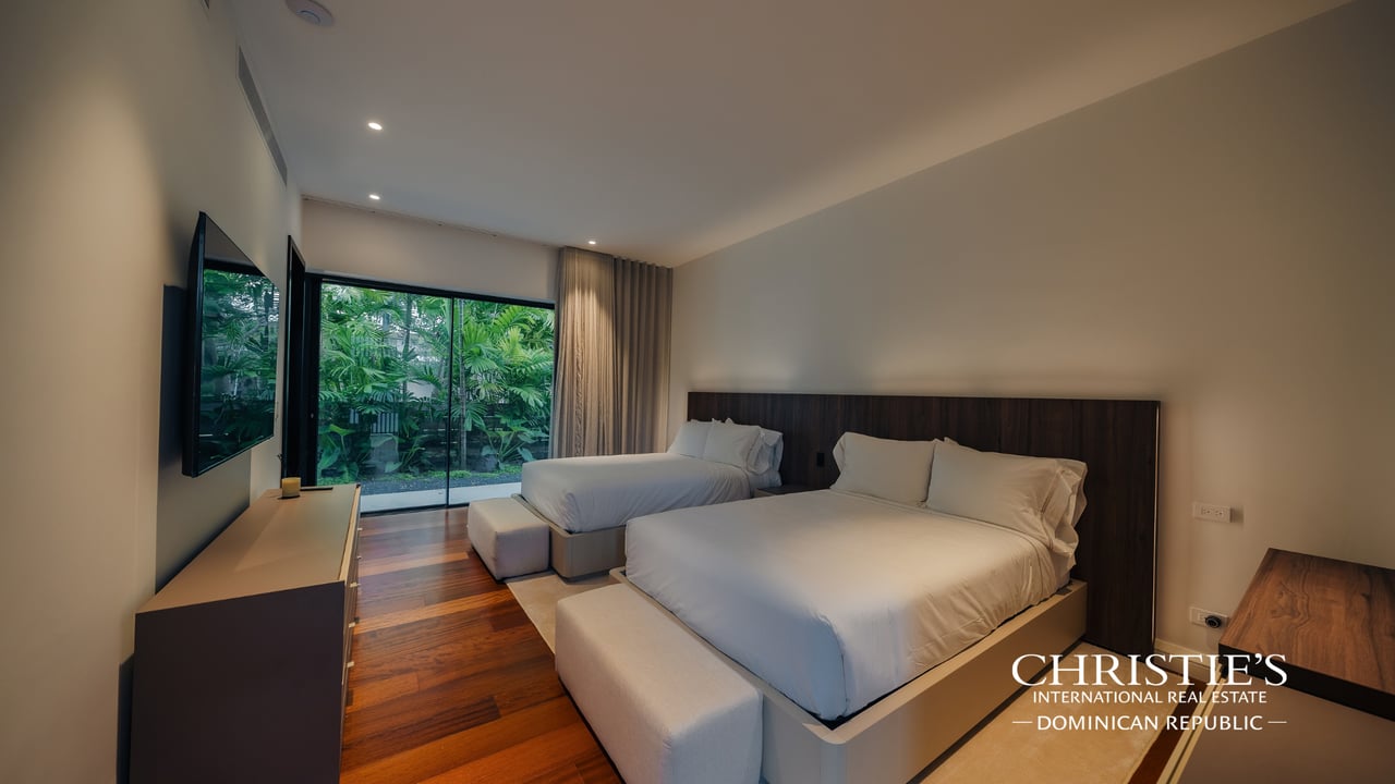 Bedroom with two beds, warm wood flooring, and glass doors overlooking tropical greenery.