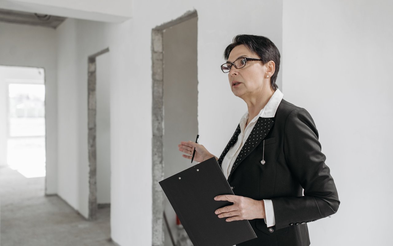 Woman with a clipboard gestures inside an unfinished building.