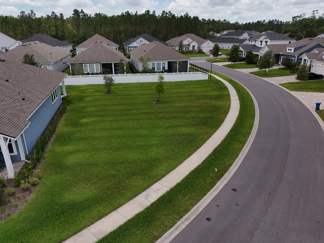 Aerial neighborhood view with detached homes, a curving street, and a large grassy vacant lot.