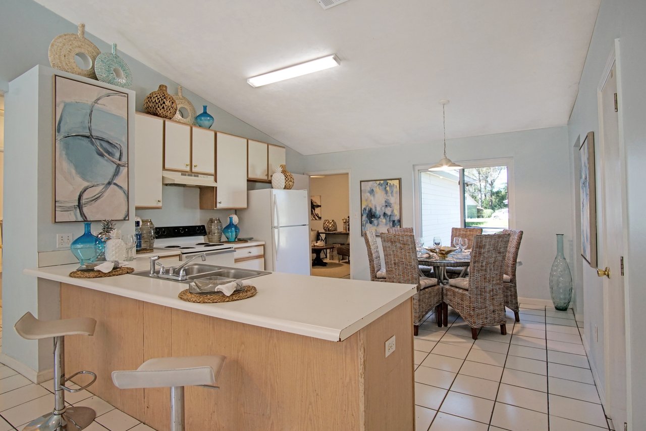 Bright kitchen with white cabinets, wicker decor, and counter stools. Adjacent dining area with a wicker table set, modern art, and pendant light. Cozy and inviting.Bright kitchen with white cabinets, wicker decor, and counter stools. Adjacent dining area with a wicker table set, modern art, and pendant light. Cozy and inviting.