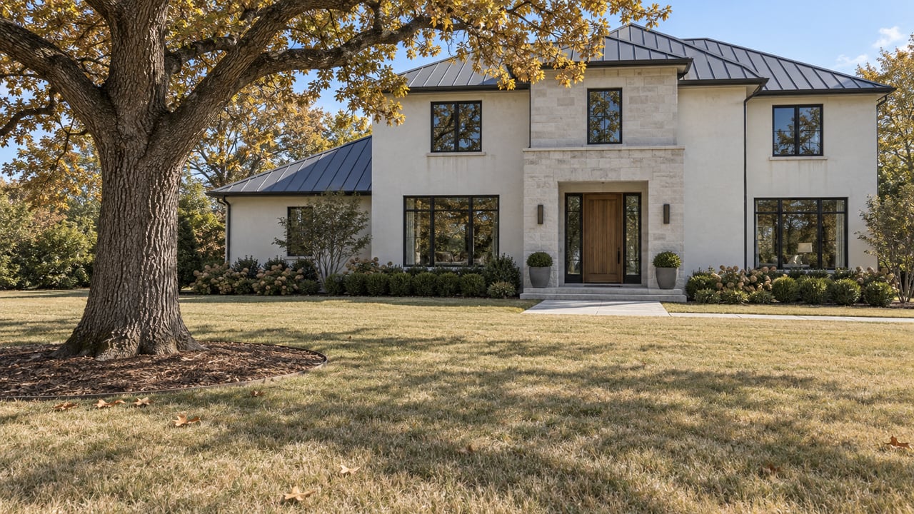 A mature bur oak stands across a broad straw-colored lawn from a pale stucco residence with dark-framed windows and a limestone entry.