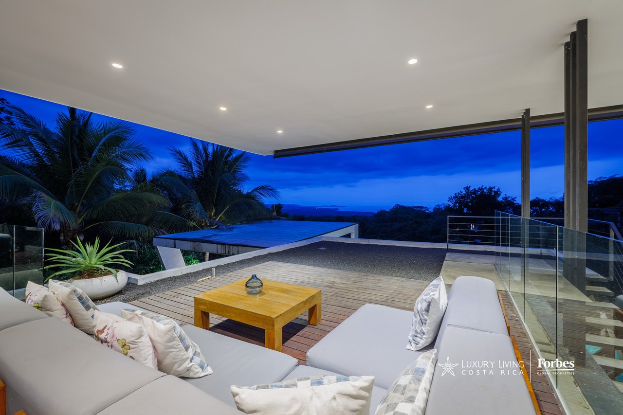 Covered terrace with white lounge seating and a wooden deck overlooking tropical greenery at dusk.