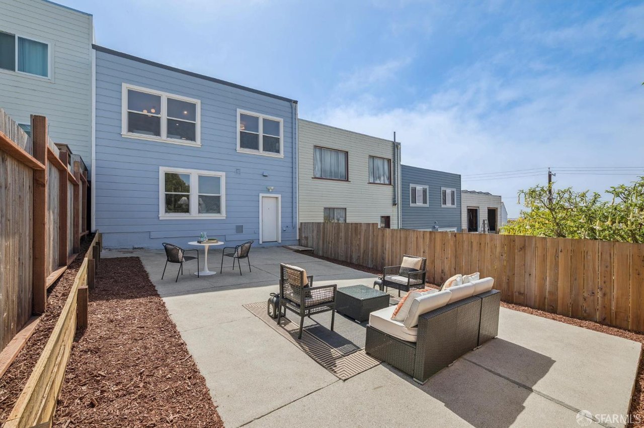 Fenced concrete patio with lounge seating and a small dining set behind a blue-sided home.
