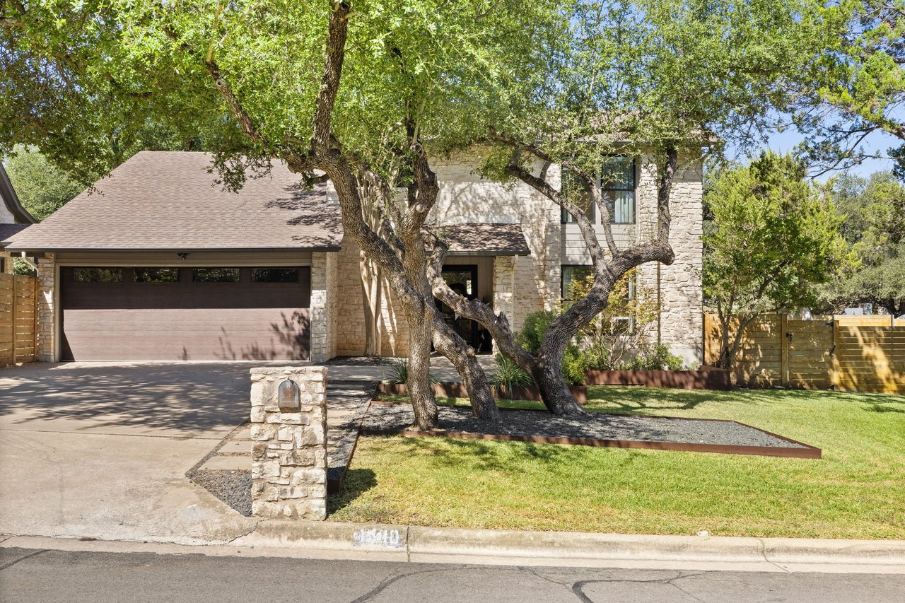 Two-story stone house with a wide garage, mature oak trees, and a stone mailbox pillar.