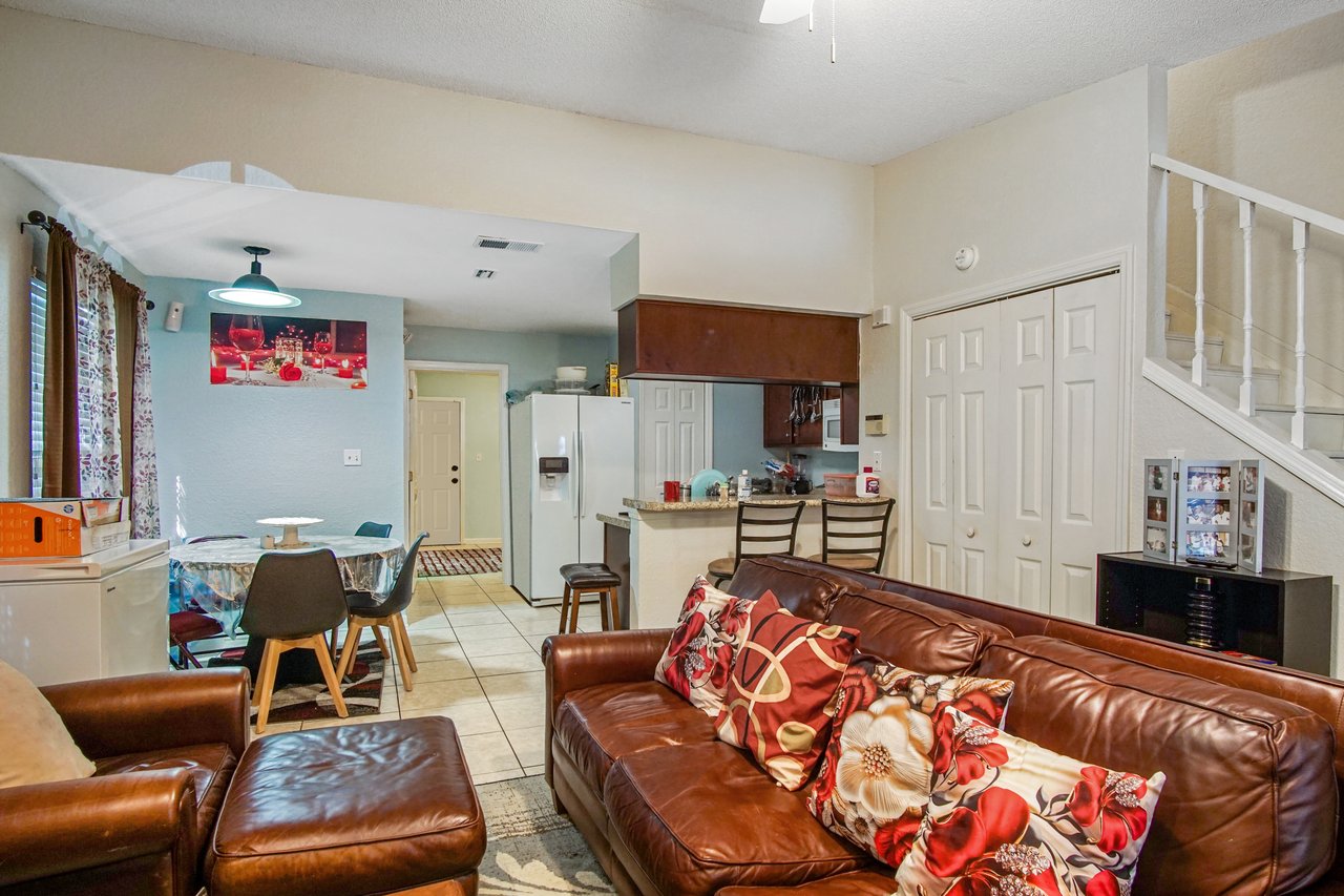 Open living area at 2506 Nugget Lane in Tallahassee, Florida, showing brown leather sofa with patterned pillows, tile flooring, and staircase with white railing. The view extends to a dining space with round table and pendant light, and a kitchen with breakfast bar, granite counters, and dark wood cabinets. A white refrigerator and double pantry doors provide storage.
