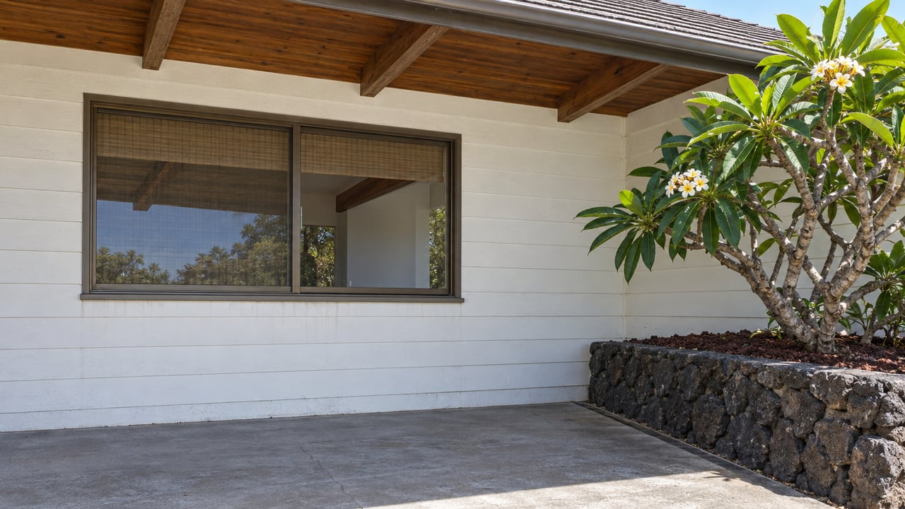Close view of a home's shaded veranda, with stained wood rafters, white siding, a bronze-framed window and a plumeria beside a lava-stone planting bed.
