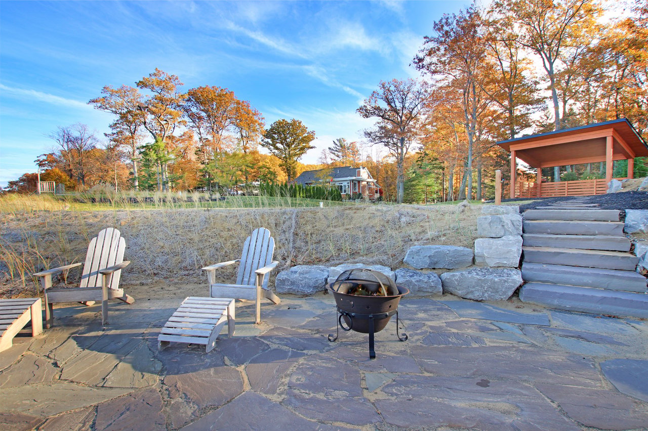 Stone patio with Adirondack chairs and fire pit, beside steps to a wooden pavilion.
