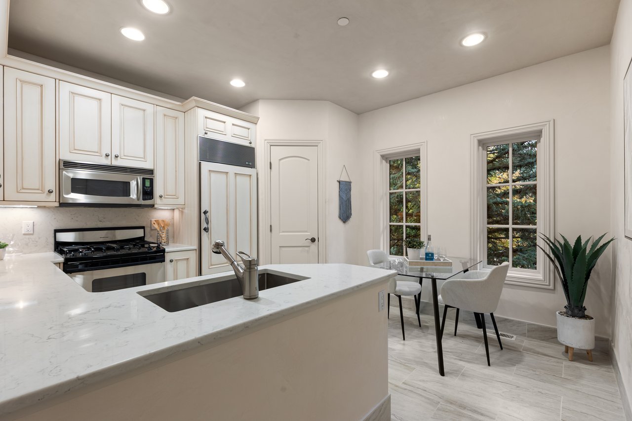 Kitchen with a large white island, stainless sink, and dining nook beside tall windows.