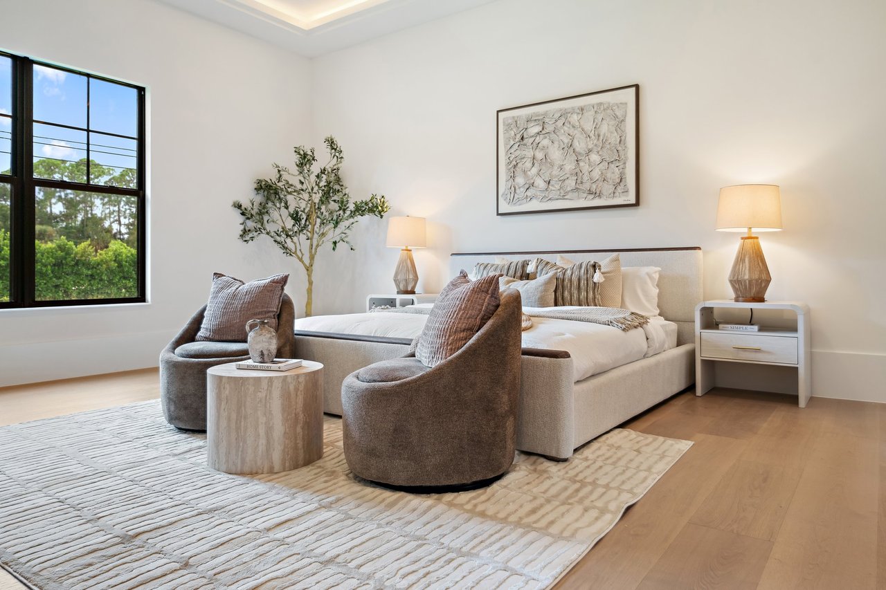 Staged bedroom with a large upholstered bed, rounded armchairs, and black-framed windows overlooking greenery.