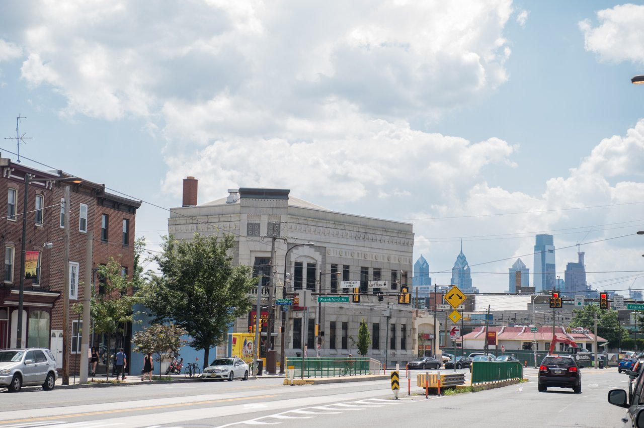 Frankford Avenue street scene with trolley tracks, a stone corner building, and Philadelphia’s skyline.