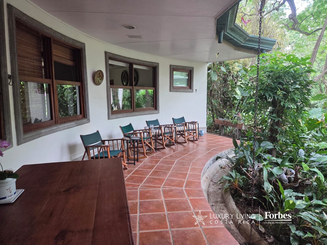 Covered villa terrace with terracotta tiles, wooden seating, and a lush garden at the edge.