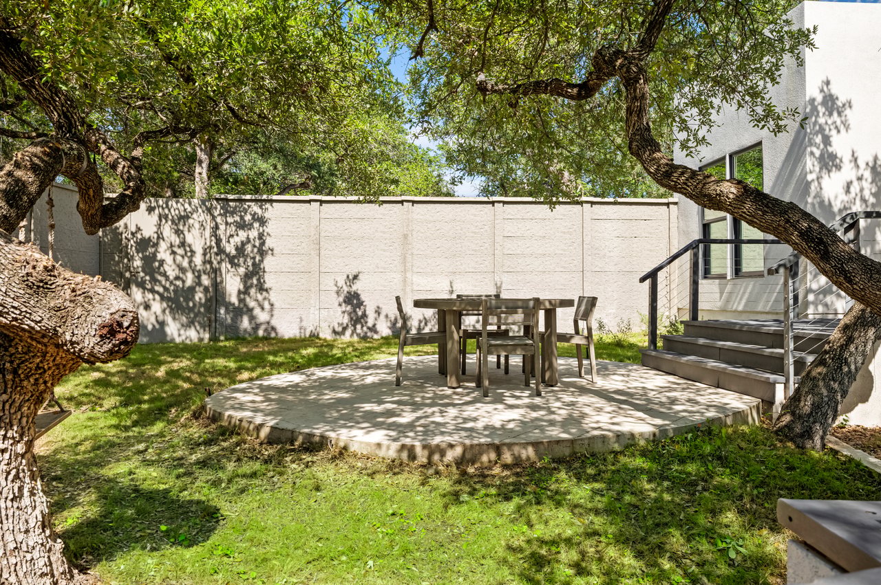 Backyard patio with outdoor dining set beneath mature trees, beside stairs to the home.