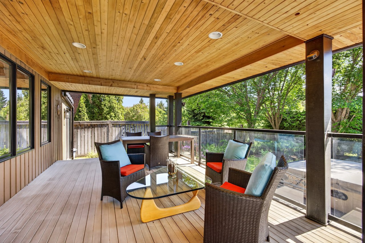 Covered deck with wicker seating, a wood ceiling, and glass railings overlooking a leafy garden.