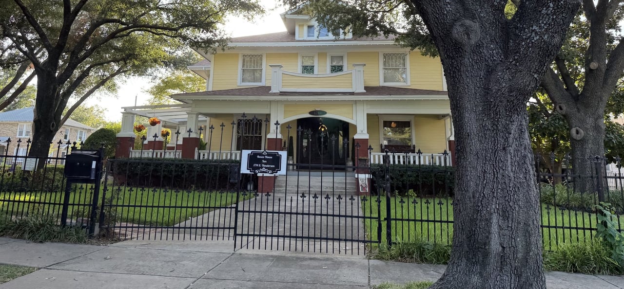 Yellow two-story historic house behind a black iron fence, framed by mature trees.