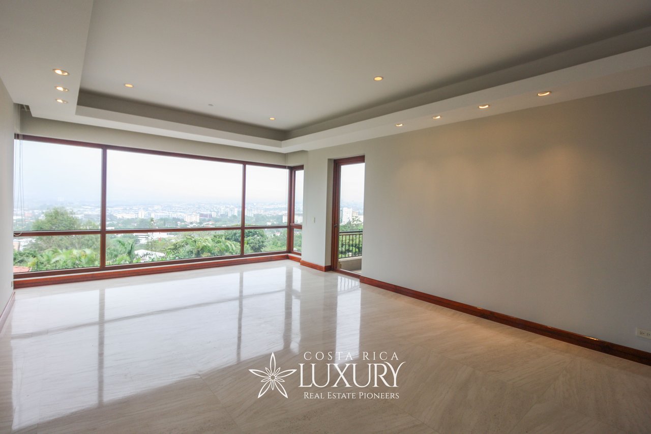 Empty living room with glossy tile floors, recessed lighting and broad windows overlooking greenery and a distant city.