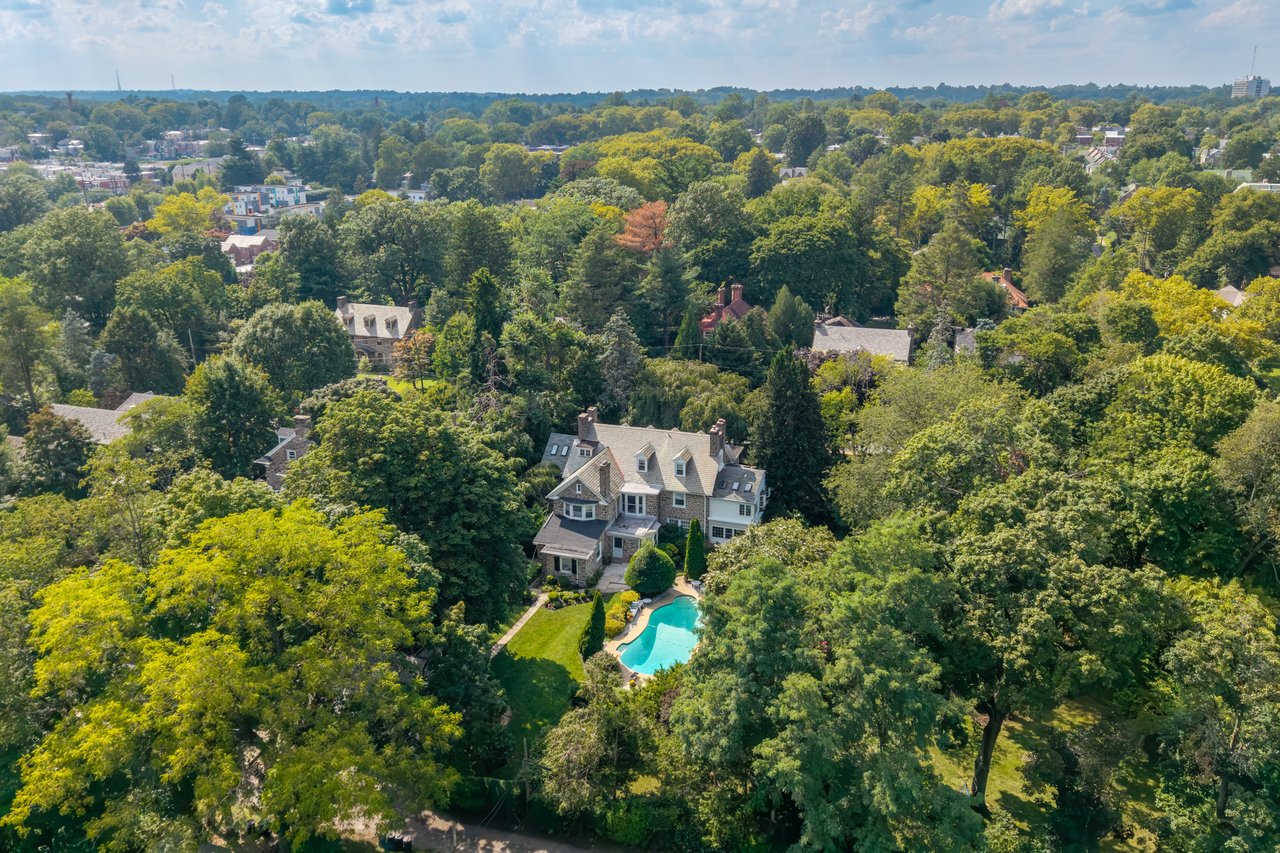 Aerial view of a stone-and-shingle home with a swimming pool amid mature trees.