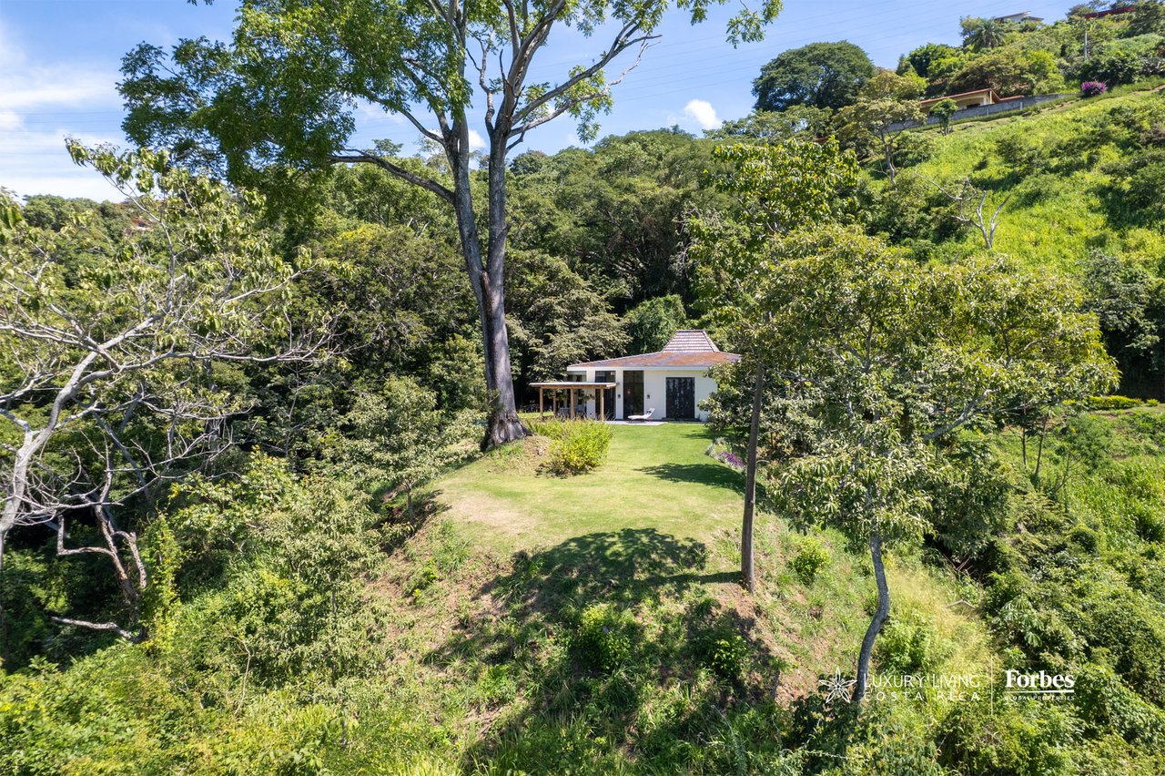 Modern home in a grassy clearing surrounded by dense tropical trees and green hillsides.