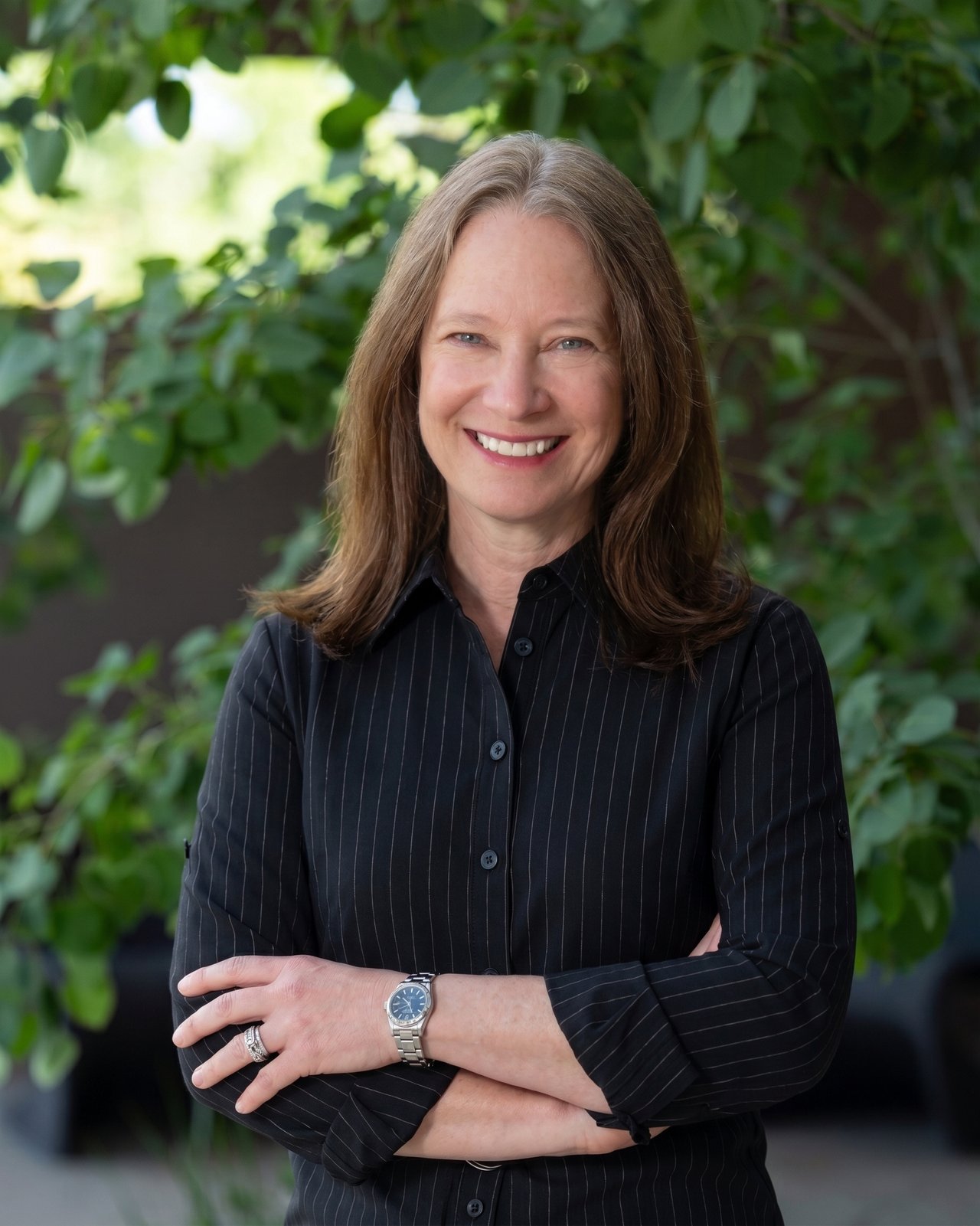 Christy Tornelli smiling with arms crossed in front of leafy greenery.